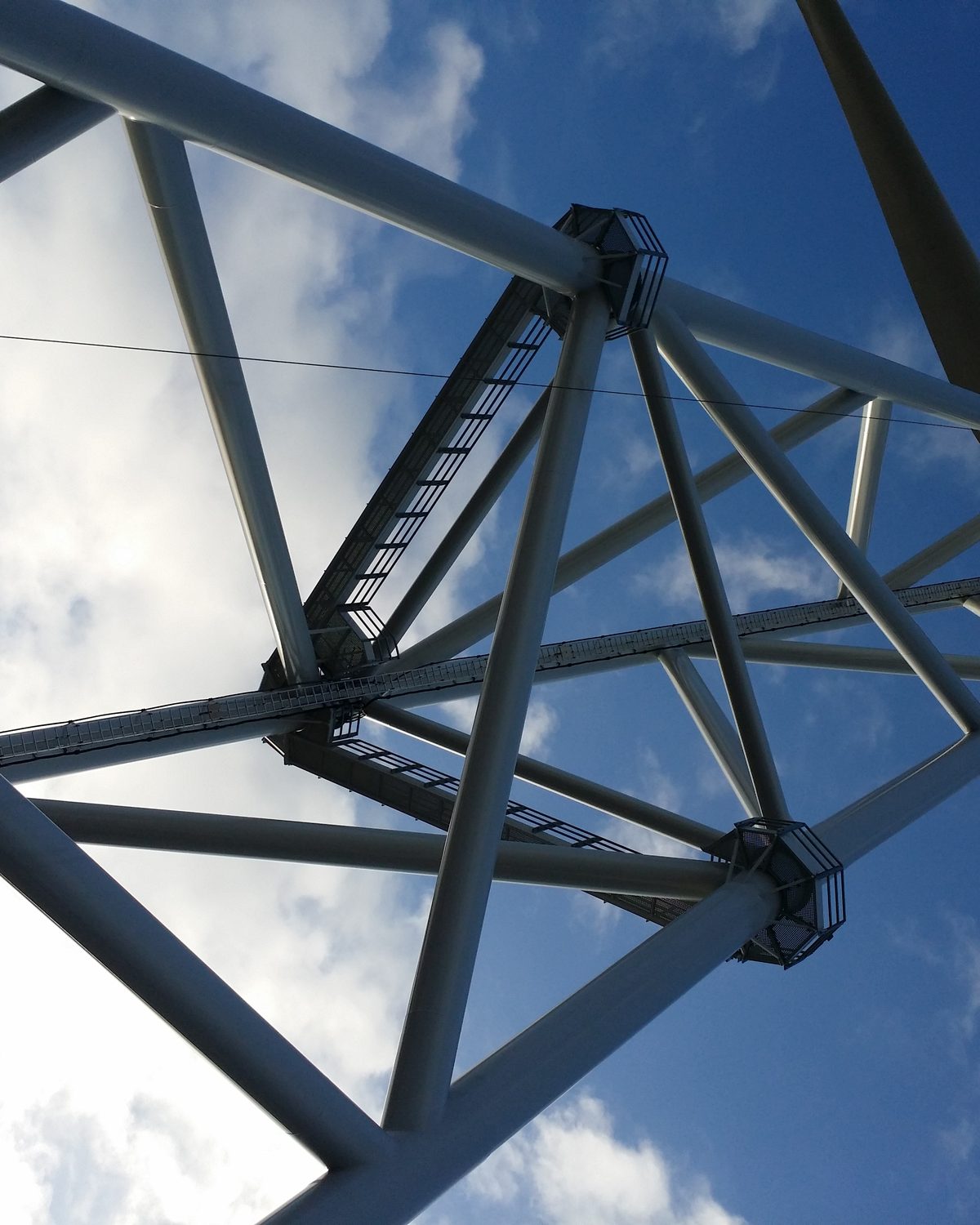 Van onderaf omhoog gezien door de vakwerkmast van een 2B-Energy-turbine, met klimladder en een rotorblad tegen de lucht.